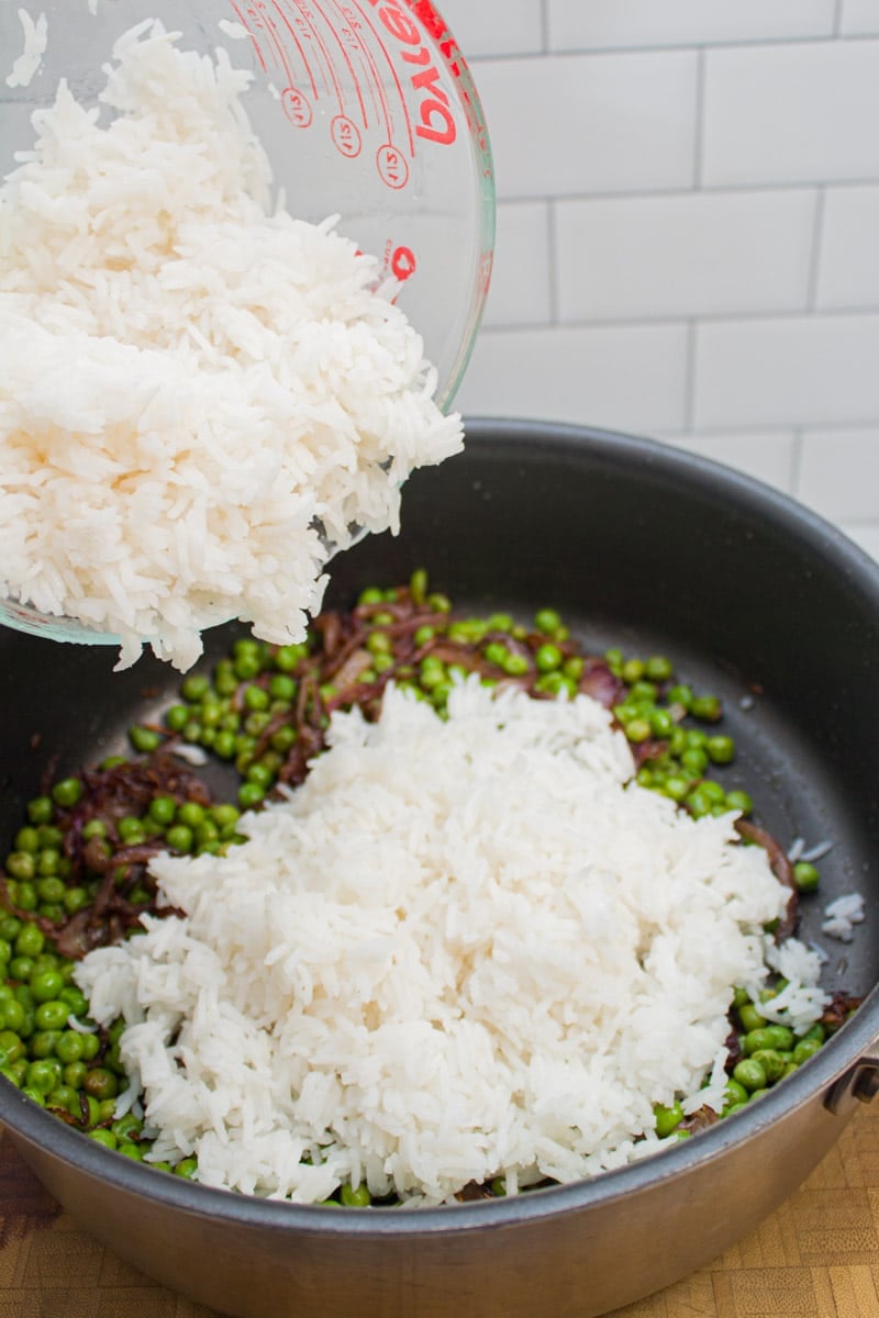 Adding basmati rice into a skillet with caramelized onions and peas.