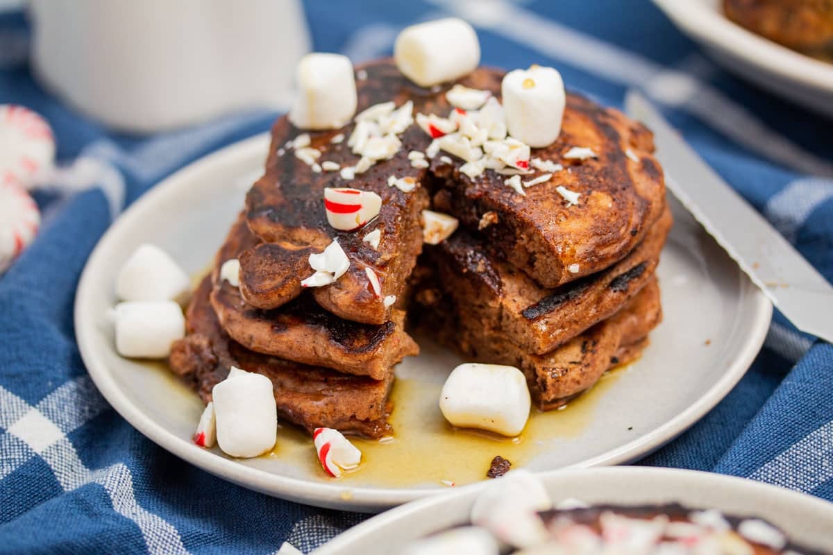 Stack of chocolate chocolate chip pancakes with peppermints cut open, garnished with mini marshmallows, and maple syrup.
