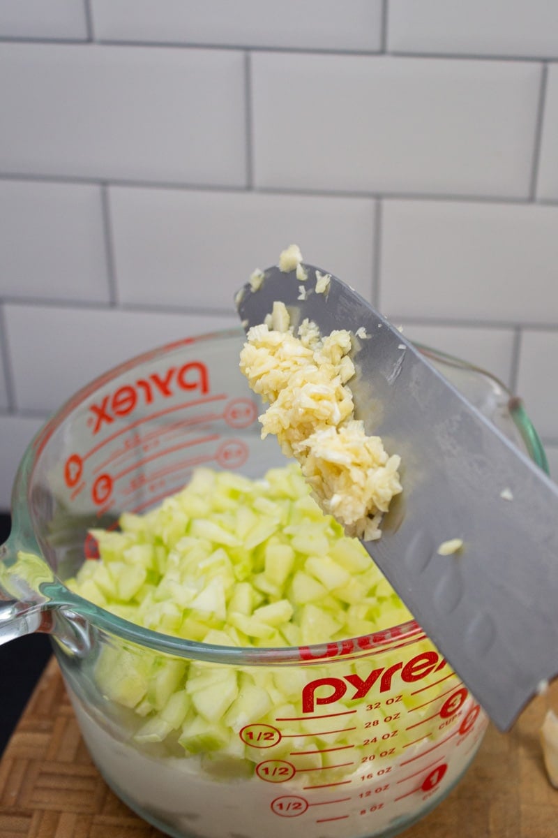 Adding garlic paste to a bowl of tzatziki ingredients.