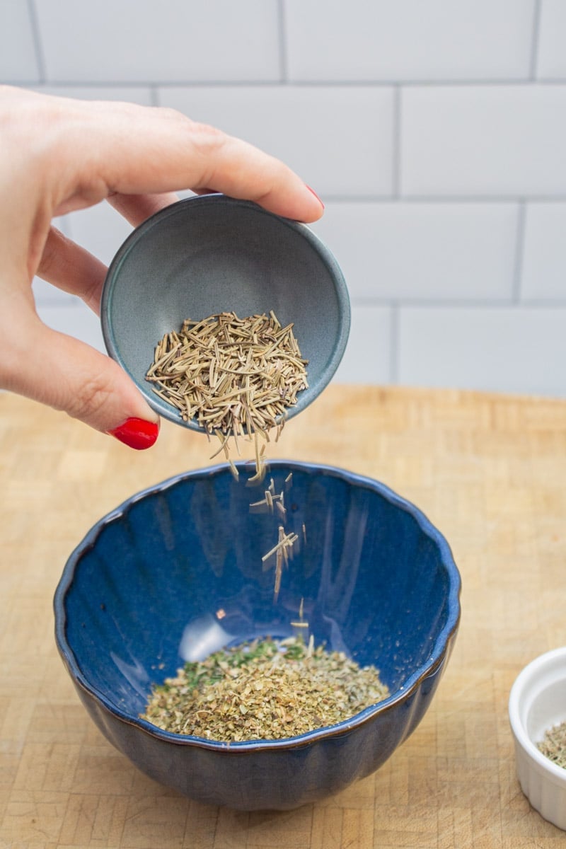 Adding dried rosemary to a bowl to make an Italian spice blend.