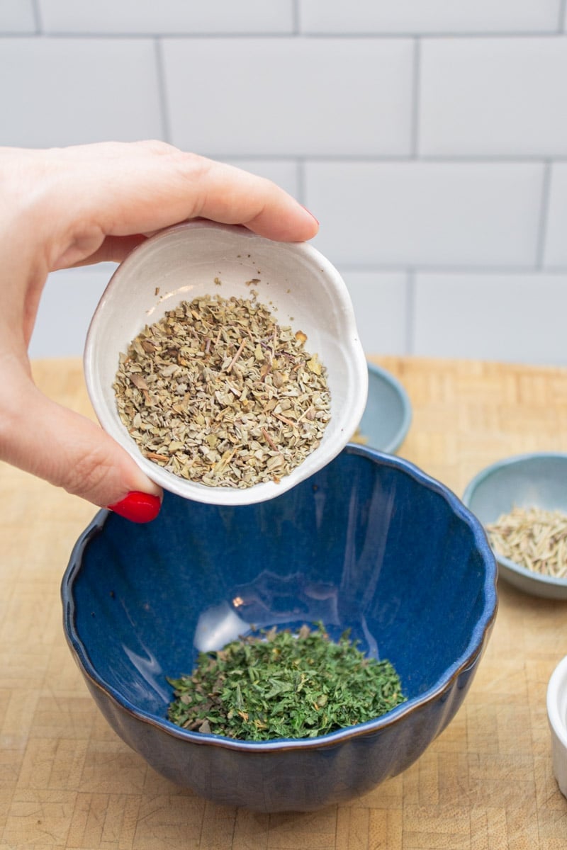 Adding dried marjoram to a bowl to make an Italian spice blend.