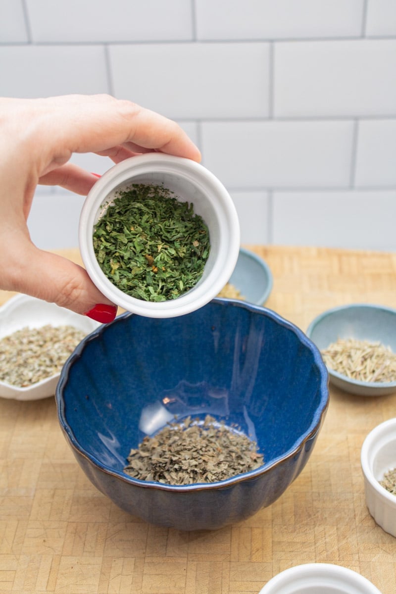 Adding dried parsley to a bowl to make an Italian spice blend.
