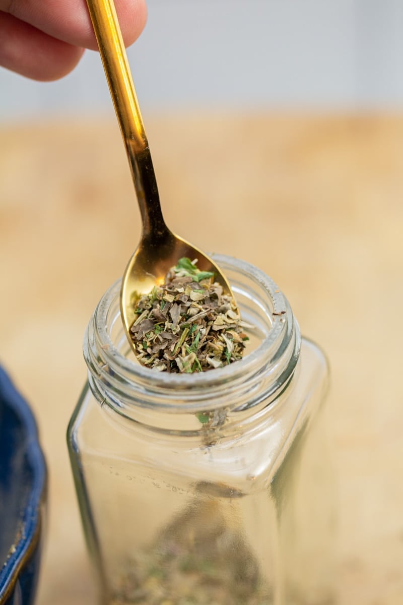 Scooping a spoonful of a homemade Italian seasoning blend into a spice jar.