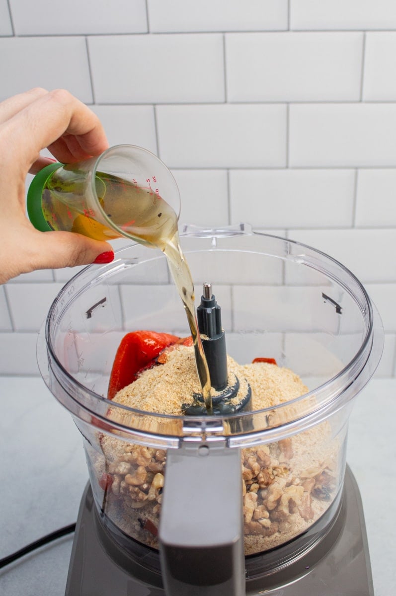 Adding liquid from a jar of roasted red peppers to a food processor with ingredients to make muhammara.