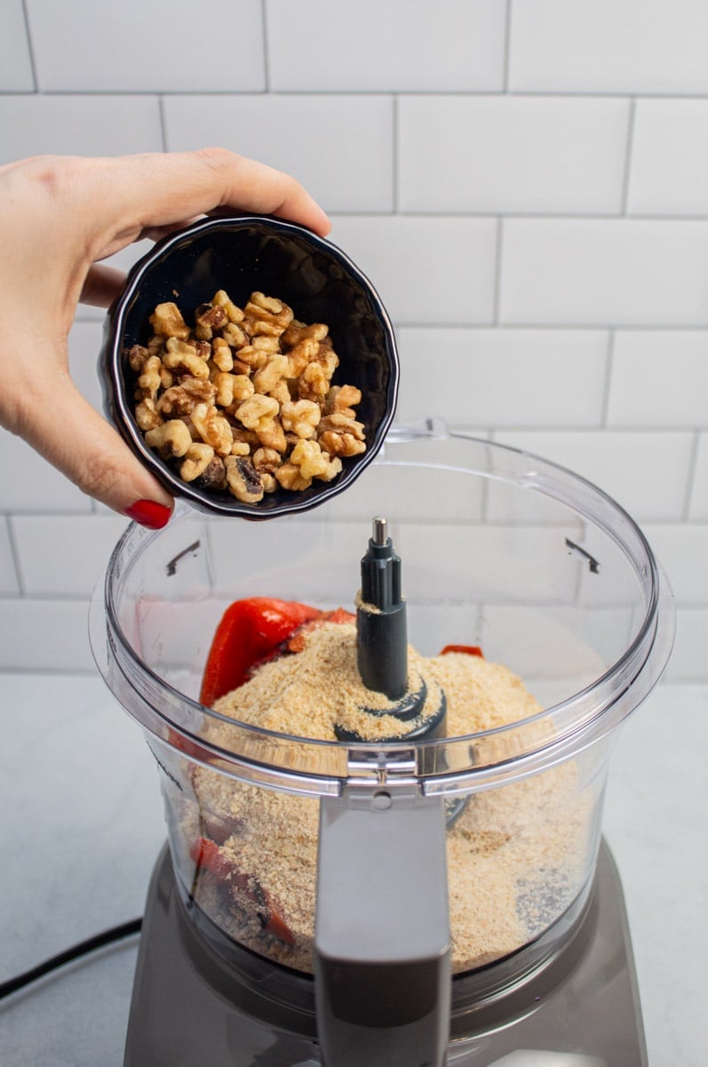 Adding walnuts to a food processor with breadcrumbs and roasted red peppers.