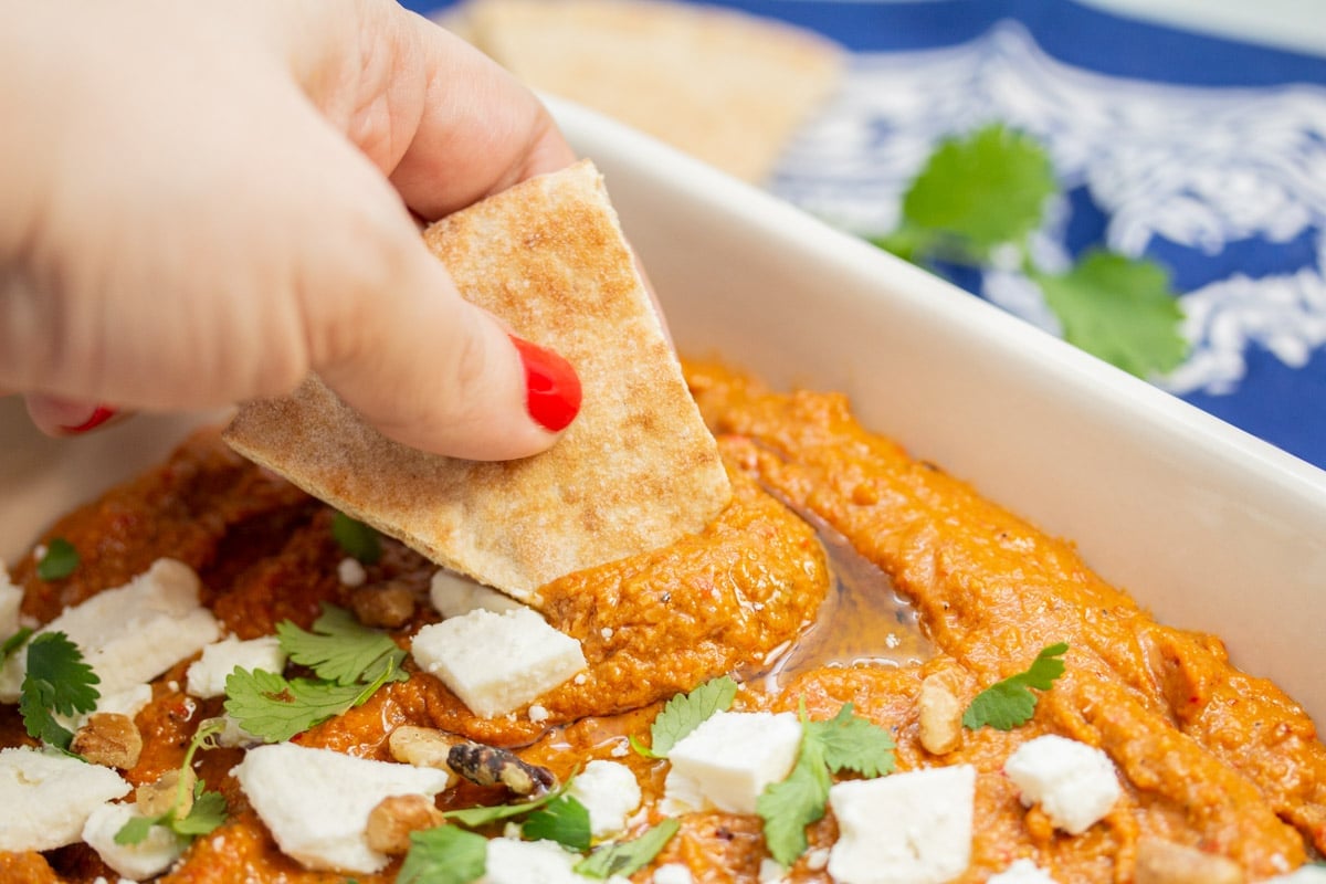 Scooping muhammara from a serving dish with a piece of pita bread.