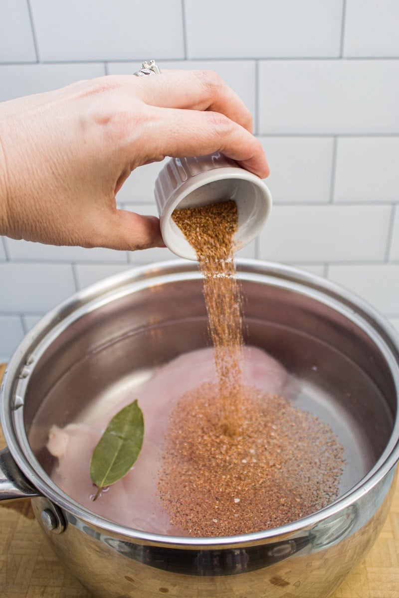 Adding seasoned salt to a saucepan with boneless skinless chicken breast, bay leaf, and water.