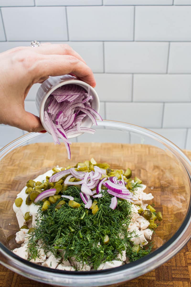 Adding sliced red onions to a bowl of dill chicken salad ingredients.