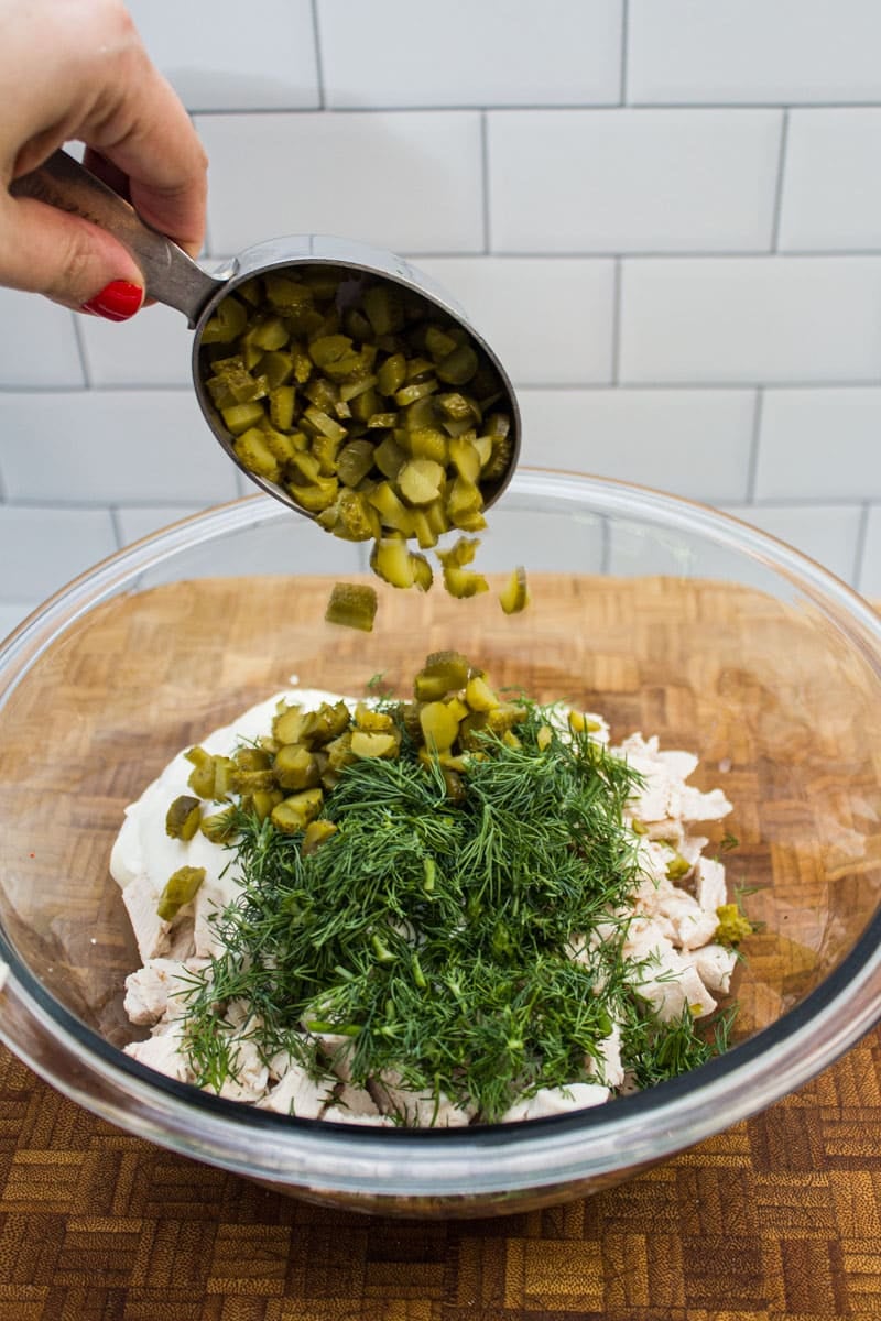 Adding chopped cornichons to a bowl of cooked chicken, chopped dill, and mayonnaise for chicken salad.